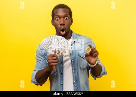Portrait de jeune homme surpris en denim chemise décontractée de la détention de monnaie dollars et physique, bitcoin dire wow avec expression, cryptocurre face choqué Banque D'Images