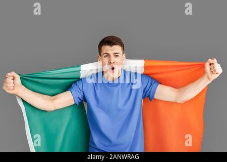 Freestyle. Jeune homme debout isolé sur gris avec drapeau irlandais soutenir équipe motivée Banque D'Images
