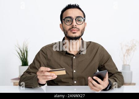 Jeune homme sérieux avec carte de crédit et votre téléphone intelligent vous regarde Banque D'Images