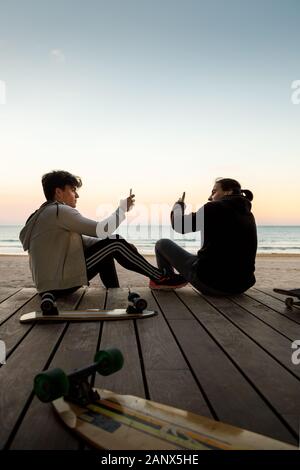 Deux jeunes garçons assis par leurs patins sur la plage pour prendre des photos avec leur téléphone cellulaire au coucher du soleil Banque D'Images