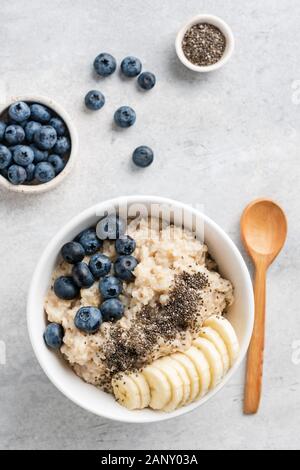 Bol de porridge d'avoine avec des tranches de banane, les graines de chia et les bleuets sur fond de béton gris, vue supérieure avec copie espace, composition verticale Banque D'Images