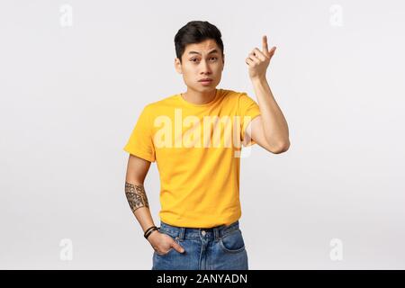 Wtf passe. Jeune homme asiatique ennuyé en t-shirt jaune, de tatouages, de l'index dans la consternation et l'irritation, soulevant les sourcils comme audience Banque D'Images