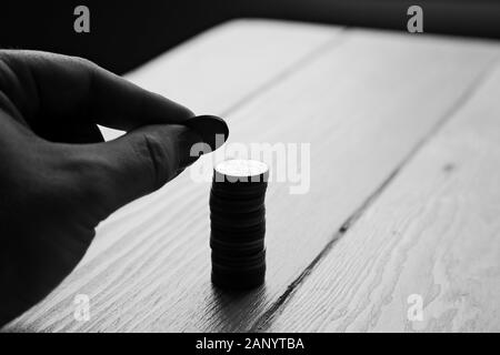 Gros plan en échelle de gris d'une personne qui met des pièces de monnaie dans une colonne sur une table en bois Banque D'Images