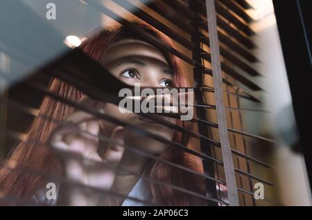 Femme asiatique regardant par des stores de fenêtres qui s'enfilent sur les voisins - jeune femme solitaire millénaires qui pleure dans le verre observer des potins et des actions à l'extérieur - introvert, espion et intrusif concepts Banque D'Images