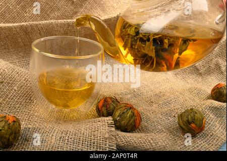 Thé aux fleurs préparé dans une théière en verre, un verre de thé et des boules de thé aux fleurs sur fond de tissu à l'Homespun rugueux. Gros plan Banque D'Images