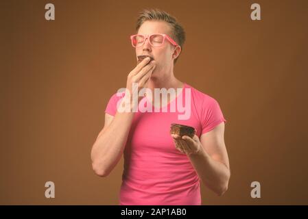 Studio shot of young handsome nerd man eating chocolate cake comme concept malpropre contre fond brun Banque D'Images