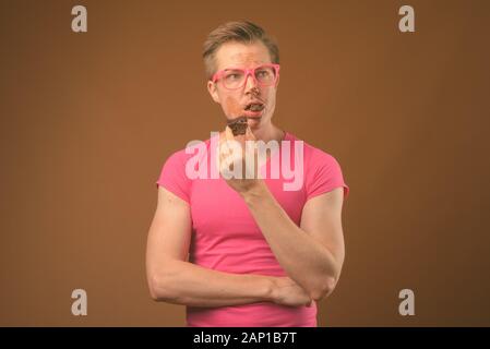 Studio shot of young handsome nerd man eating chocolate cake comme concept malpropre contre fond brun Banque D'Images