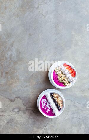 Des bols de smoothie pour petit déjeuner sains, surmontés de pitaya, de noix de cajou, de flocons de noix de coco, de muesli et de fraise sur fond gris. Espace de copie. Banque D'Images