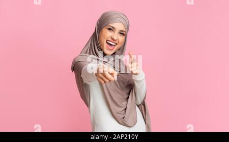 Femme arabe positive montrer du doigt ce qui pose l'appareil photo sur fond rose Banque D'Images