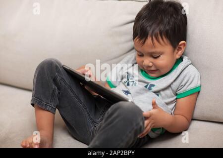 Captivés par l'app qu'il joue avec, un mignon petit garçon est allongé sur un canapé et tenant une tablette sur ses genoux. Banque D'Images