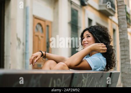 Smiling girl avec des cheveux bouclés et assis sur un banc à l'écart avec une attitude réfléchie Banque D'Images