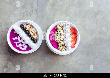 Des bols de smoothie pour petit déjeuner sains, surmontés de pitaya, de noix de cajou, de flocons de noix de coco, de muesli et de fraise sur fond gris. Espace de copie. Banque D'Images