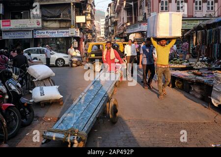 Un porteur pousse son chariot à travers la zone occupée de bazar de Bhendi à Mumbai, en Inde, un autre transportant des marchandises sur sa tête Banque D'Images