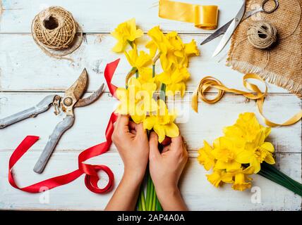 Vue de dessus des mains fleuristes faisant bouquet de fleurs avec narcisse et ruban décoratif. Fleuriste vintage et fabrication de bouquet au printemps. Banque D'Images