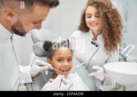 Dentiste expérimenté en uniforme médical blanc et gants examinant la cavité orale de la jolie fille africaine avec l'aide d'une assistante féminine. Médecin utilisant une sonde dentaire et un miroir au travail. Banque D'Images