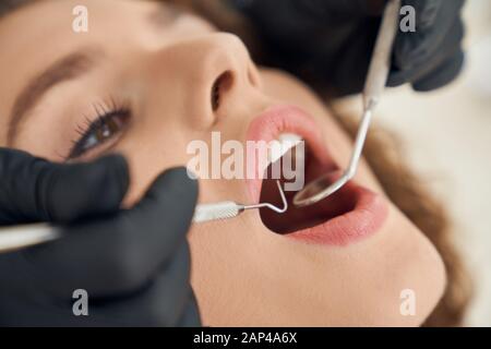 Gros plan sur la belle jeune fille assise dans une chaise dentaire avec bouche ouverte alors que le dentiste en gants noirs à l'aide de la sonde et du miroir pour le contrôle oral. Concept de diagnostic régulier. Banque D'Images