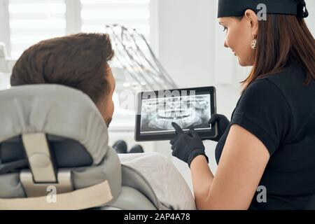 Vue latérale d'un dentiste féminin confiant en uniforme médical noir, en gardant le comprimé et en montrant une image radiographique des dents au client masculin assis sur une chaise dentaire. Concept de médecine et de soins de santé Banque D'Images
