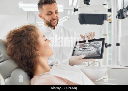 Vue latérale d'une belle fille avec des cheveux frisés assis dans une chaise dentaire et regardant l'appareil électronique qui tient un médecin de beau sexe masculin. Dentiste professionnel montrant des photos de rayons X de dents au client Banque D'Images