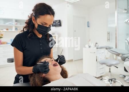 Dentiste professionnel de sexe féminin en uniforme médical noir, masque et gants en caoutchouc examinant la cavité buccale du patient à l'aide d'une sonde dentaire et d'un miroir. Concept de dentisterie et de traitement de la santé. Banque D'Images