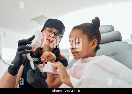 Dentiste professionnel de sexe féminin en uniforme médical noir, gants et lunettes montrant comment brosser les dents sur le modèle de mâchoire dentaire. Petite dame africaine assise dans une chaise dentaire et regardant attentivement Banque D'Images