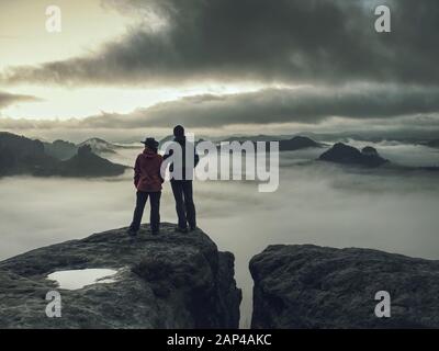 Les randonneurs la femme et l'homme dans les vêtements sombres sur plus haute montagne et regarder le lever du soleil dans l'obscurité. Sur le bord du sentier rocheux au-dessus de la vallée brumeuse. Tourist couple enj Banque D'Images