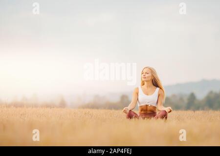 Jeune fille en chemise blanche, vêtements de sport avec cheveux longs Blond médite le yoga dans le champ lever du soleil. Concept de relaxation Banque D'Images