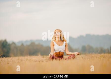 Jeune fille en chemise blanche, vêtements de sport avec cheveux longs Blond médite le yoga dans le champ lever du soleil. Concept de relaxation Banque D'Images