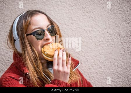 Belle jeune fille mignonne dans des vêtements à la mode bénéficie d'hamburger de restauration rapide en position debout en face d'un mur Banque D'Images