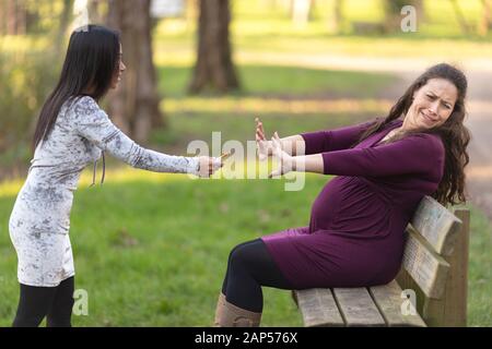 Femme enceinte, assis sur le banc dans le parc et en rejetant la cigarette offerte par une autre femme, avec des gestes d'arrêt. Éviter les mauvaises habitudes et healt Banque D'Images