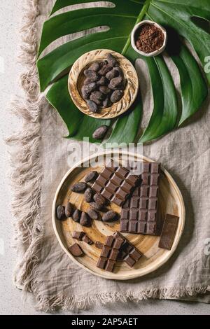 Barre de chocolat noir et au lait ensemble et hachées dans une plaque en céramique avec les fèves de cacao et poudre de cacao sur feuilles monstera exotiques sur tissu en lin gris comme back Banque D'Images
