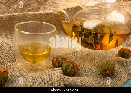 Thé aux fleurs préparé dans une théière en verre, un verre de thé et des boules de thé aux fleurs sur fond de tissu à l'Homespun rugueux. Gros plan Banque D'Images