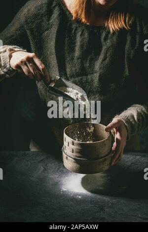 Procédé de fabrication de pâte à pain fait maison. Femme mains tamise la farine du vintage sur tamis de cuisine sombre. Tableau noir avec de la farine. Accueil la cuisson du pain. Des Photo s Banque D'Images