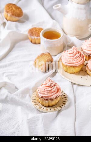 Petits gâteaux faits maison avec crème au beurre rose et flocons de noix de coco servi avec théière en céramique, tasse de thé blanc sur nappe pliée. Banque D'Images