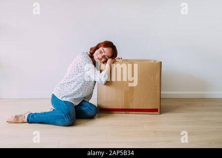 Jolie fille en train de se déplacer dans le nouvel appartement. Assis sur le sol avec boîte en carton dans une pièce vide, souriant. Rêvant du jour, fond de mur blanc. Banque D'Images