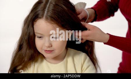 Maman braid le tresse de sa fille avec de longs cheveux. Séparément sur un fond blanc. Une fille de six ans joue, et maman plie sa queue de porc. Banque D'Images