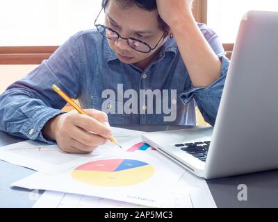 Homme d'affaires asiatique portant une chemise bleue et des lunettes tenant le crayon et pensant à travailler avec un ordinateur portable sur la table près de la fenêtre au bureau. Banque D'Images