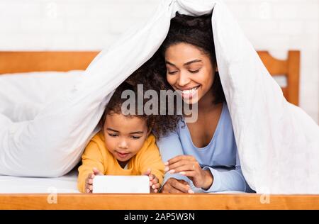 Mère Et Fille Africaines Utilisant Le Téléphone Mobile Allongé Dans Le Lit Banque D'Images