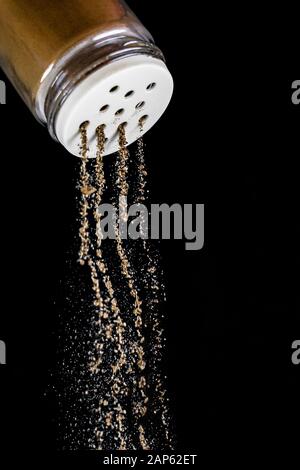 Le poivre noir est sorti de l'agitateur à poivre sur un fond noir isolé. Le processus d'assaisonnement s'arrête à temps. Agitateur à sel de verre avec St blanc Banque D'Images