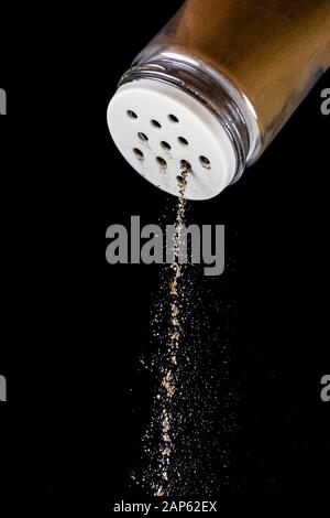 Le poivre noir est sorti de l'agitateur à poivre sur un fond noir isolé. Le processus d'assaisonnement s'arrête à temps. Agitateur à sel de verre avec St blanc Banque D'Images