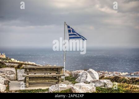 Drapeau grec flamant une journée venteuse à côté de la table de pique-nique en bois. Ermoupolis sur l'île de Syros, à l'extrême droite. Banque D'Images