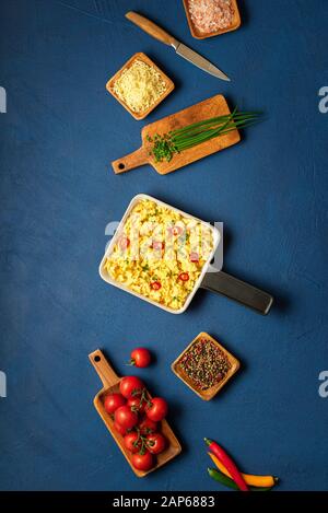 Œufs brouillés, fraîchement cuits, dans une poêle et des ingrédients en céramique, fromage et tomates, sur une table bleue. Concept de petit-déjeuner maison. Omelette et fromage. Banque D'Images