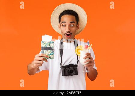 Amusant gars afro-américain heureux enfin en vacances, aller à l'étranger, tenant un cocktail et passeport avec deux billets, regardant étonnés et joyeux Banque D'Images