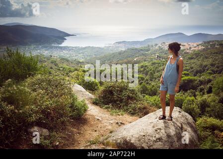 Jeune femme en vacances d'été en denim dungaree portant des shorts et sandales admiring panorama à partir de la colline qui surplombe bay en Toscane, Italie Banque D'Images