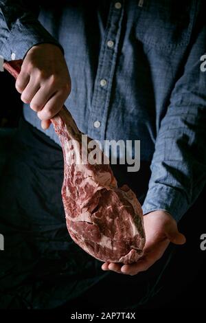 Man's hands holding non cuites brutes tomahawk boeuf Black Angus Steak sur les os sur fond sombre. Style rustique Banque D'Images