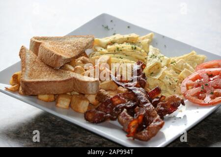 des tranches de bacon de porc croustillantes et bien cuites frites ou grillées avec des hash browns et des toasts avec des tomates sur plaque dans une vue rapprochée pour le déjeuner ou le dîner du petit déjeuner Banque D'Images