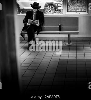 Un homme élégant portant un costume et un chapeau lit un journal assis dans la salle d'attente alors qu'il attend un train, Kent, Angleterre Royaume-Uni Banque D'Images