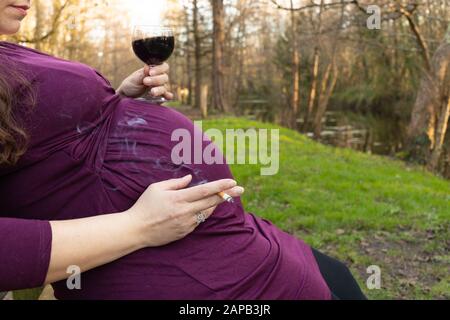 Femme enceinte, assise sur un banc, de boire du vin rouge et cigarette dans un parc. Banque D'Images