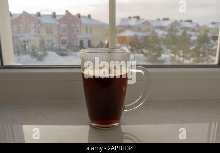 Grande tasse de verre transparent de la boisson chaude du matin - café ou thé sur une fenêtre avec de belles maisons colorées d'hiver comme arrière-plan . Tasse à café sur neige Banque D'Images