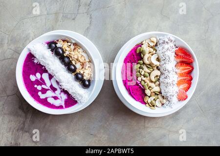 Des bols de smoothie pour petit déjeuner sains, surmontés de pitaya, de noix de cajou, de flocons de noix de coco, de muesli et de fraise sur fond gris. Espace de copie. Banque D'Images