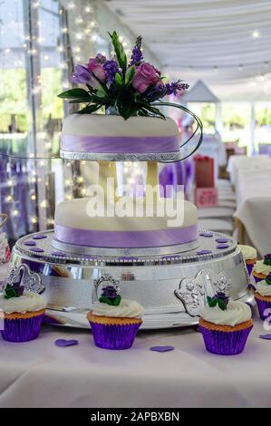 Gâteau de mariage violet pâle et blanc sur une table avec des cupcakes. Banque D'Images